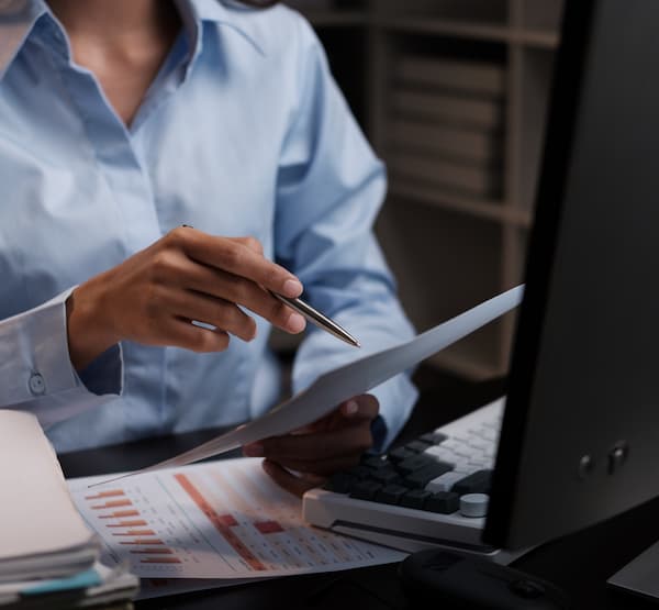 Man at a desk in front of a computer and papers, writing something on a pile of papers
