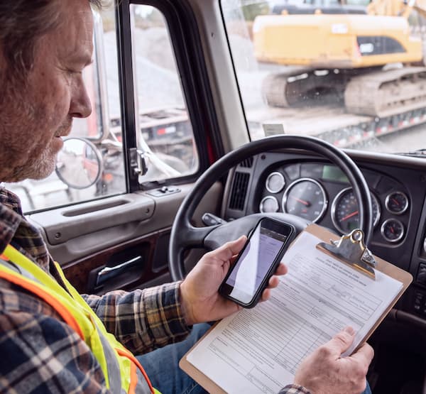 Truck driver sitting in a parked truck reviewing a document on a clipboard and their smartphone